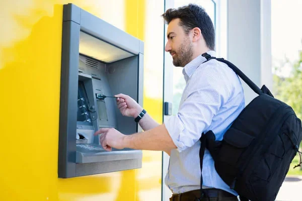 Mężczyzna korzysta z bankomatu obok żółtej ściany, z plecakiem na ramieniu.